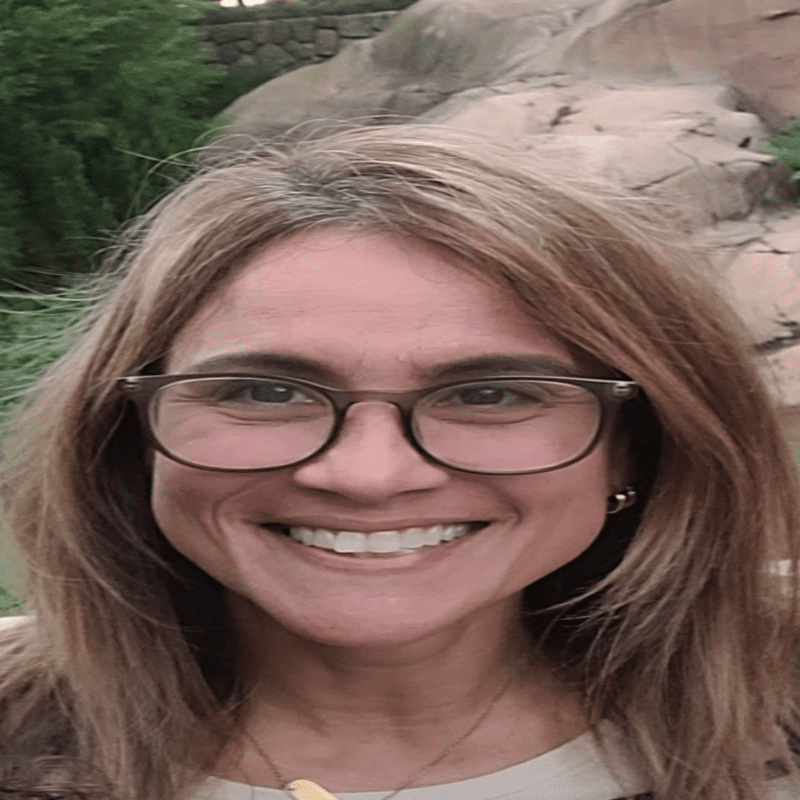 A smiling woman with glasses outdoors near rocks and greenery.